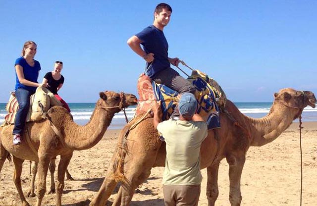Three students ride camels on a beach in Morocco