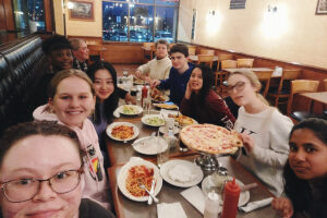 Students smile at a table filled with pizza, pasta, and other dishes.