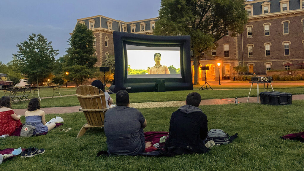 Students sit on the grass of the Quad, watching a movie on a large screen in front of Pardee Hall.