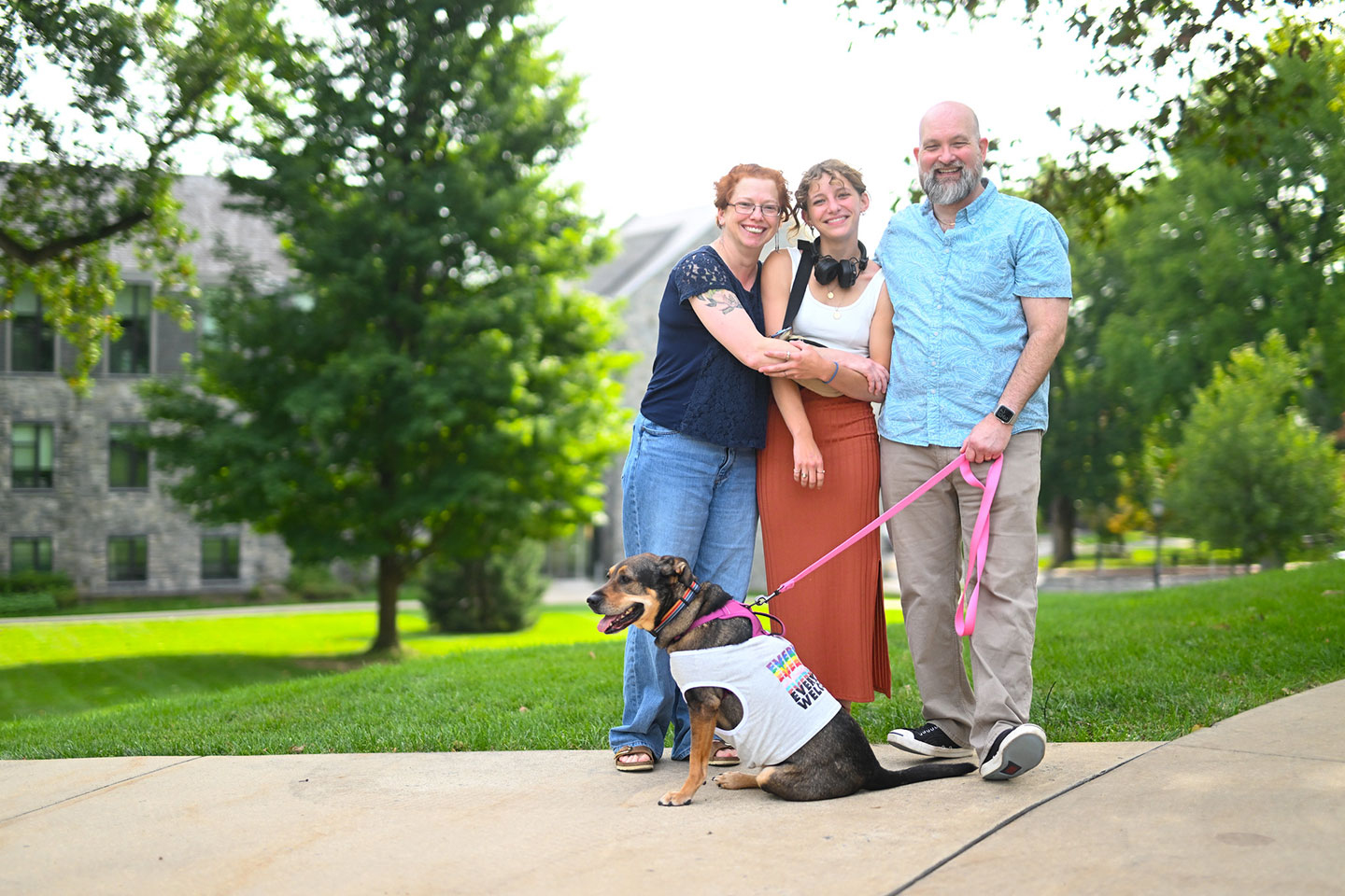Mom and dad are hugging their daughter while dad is holding the leash for their dog. The dog is wearing a t-shirt. 