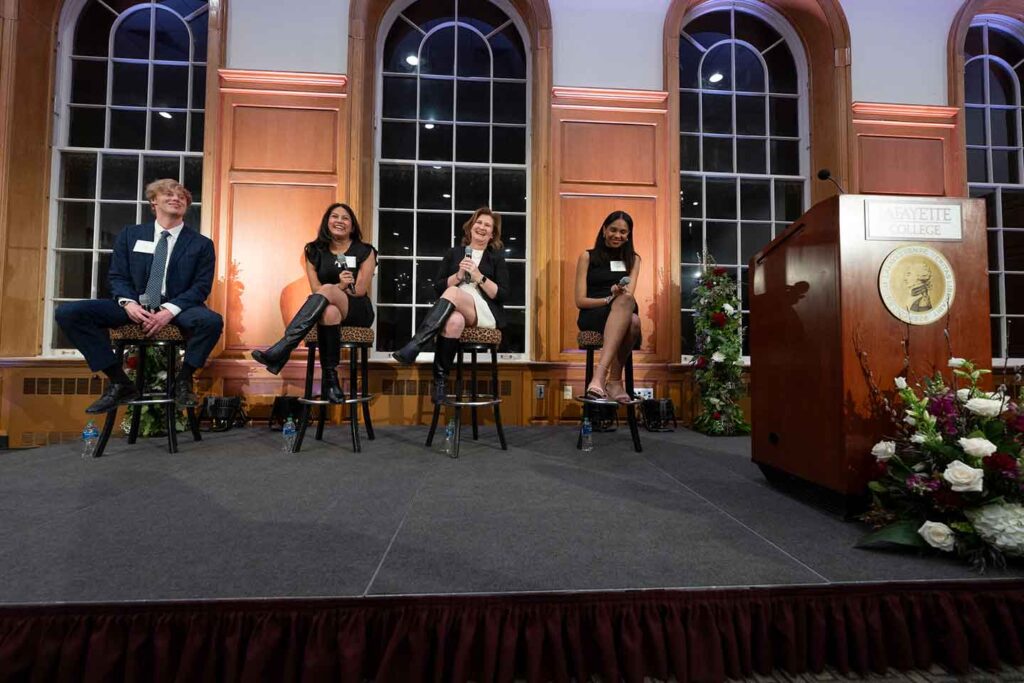 Three students and President Hurd are seated on leopard stools on a podium.