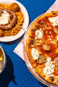 A sausage and cheese topped pizza and a plate of spaghetti and sausage are shown on a blue tablecloth
