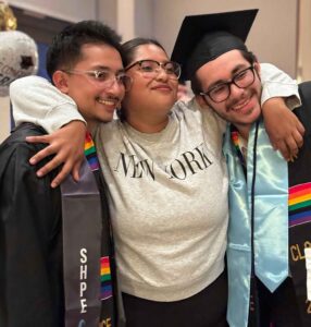 Three students stand and smile at the camera. Two of the students are wearing graduation gowns and stoles. The student on the right is wearing a graduation cap.