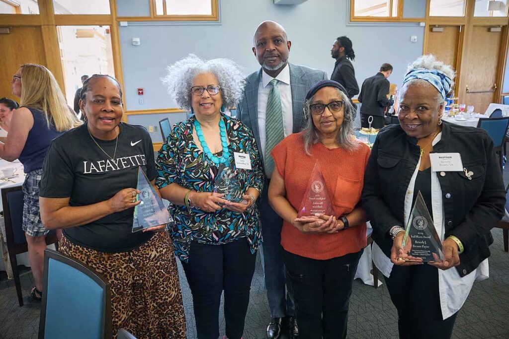 A group of four alumnae are holding glass awards. They are smiling at the camera. Ernest Jeffries, vice president of inclusion is standing behind them.
