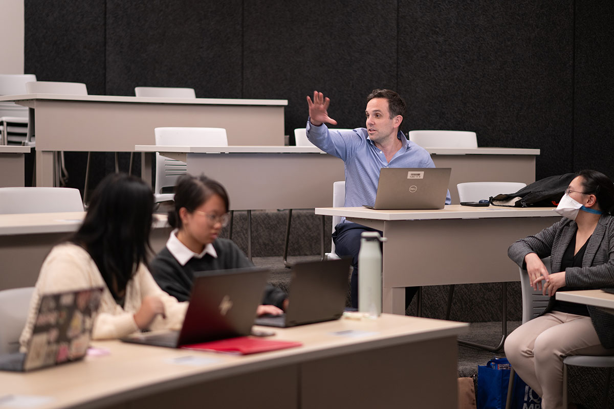 Prof. Dylan Groves gestures to a room of students. 