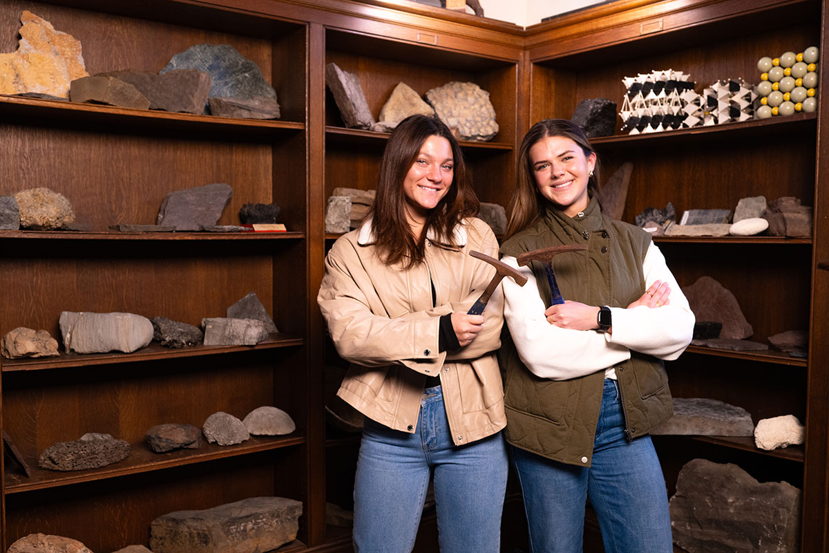 Ellie Spencer and Kate Marrs stand back to back in front of a bookshelf full of rocks holding hammers. 