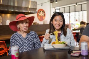 Two women sit in front of Mister Lee's Noodles at the Easton Public Market. One uses chopsticks to hold up ramen noodles