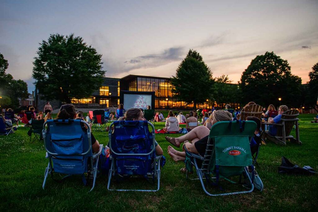 Students, staff, and neighbors sit in chairs on the quad to watch a movie set up on an inflatable screen