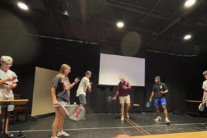 Students stand in a black box theater holding tennis rackets, performing a self written play.