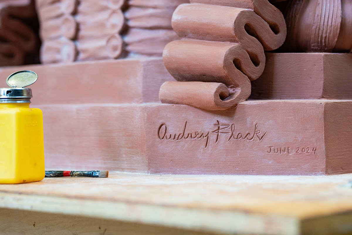 A clay scroll is shown with the artist signature (Audrey Flack) carved in the clay. A yellow jar of paint sits nearby.