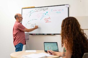 Prof. Chris Ruebeck standing at a whiteboard and Mita Crane '26 sitting at a table, discussing a research project impacting Ruebeck's Industrial Organization course