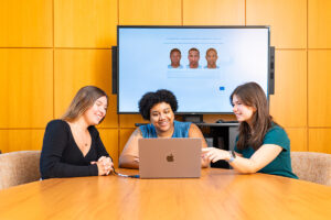Deja Simon-Jennings, Charlotte Bowler ’27 and Erin Rapuano ’27 sit at a long desk and observe a computer, with a screen turned on behind them.