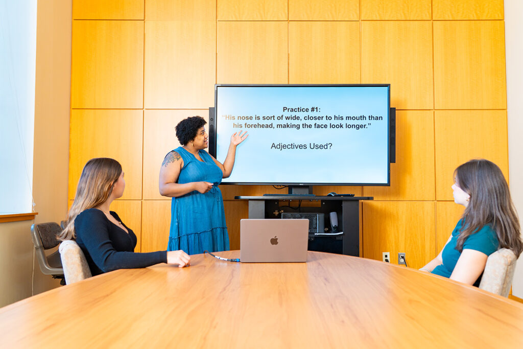 Charlotte Bowler ’27 and Erin Rapuano ’27 sit at a long desk and observe a computer, with a screen turned on behind them. Deja Simon-Jennings teaches.