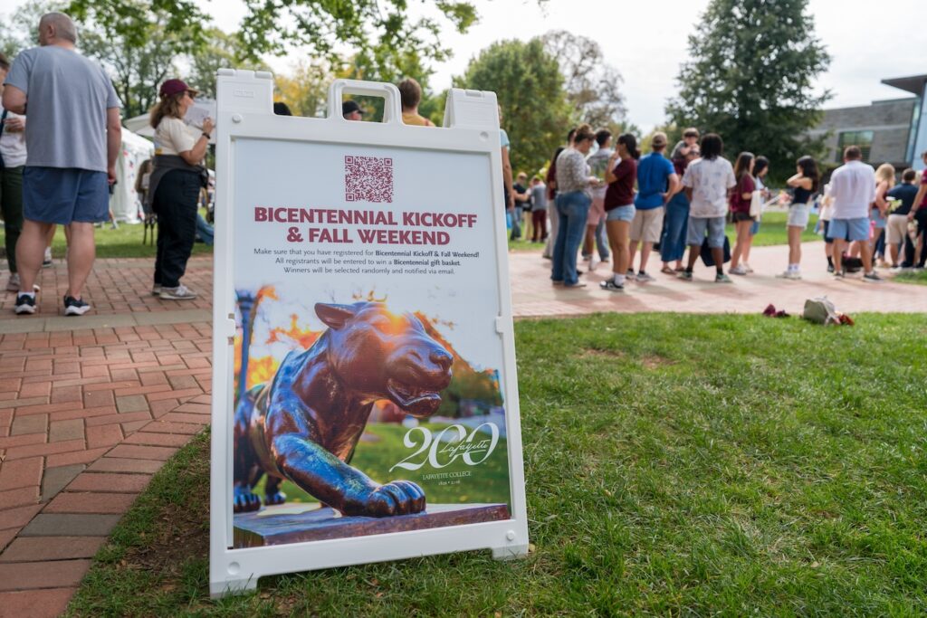 An A-frame sign sits on the Quad with info about the Bicentennial Kickoff and Fall Weekend. People are seen walking through campus in the background.