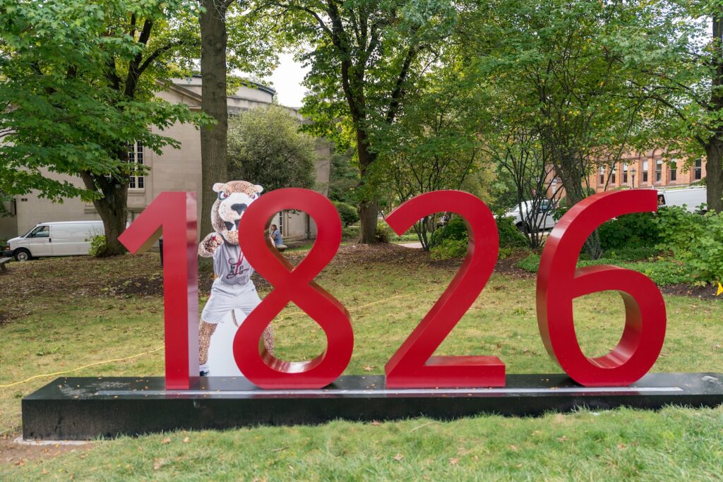 A maroon 1826 sign stands on the Quad with an image of the Leopard behind it.