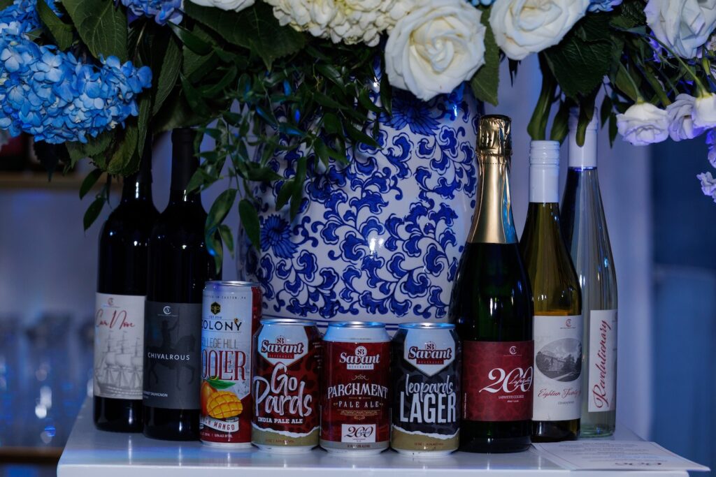 Bottles of five Lafayette-themed wines and four Lafayette-themed canned beer and wine coolers are shown with a vase of flowers in the background.