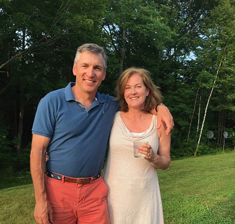Brad '87 and Kristen '86 Lewis are standing outside. Kristen is holding a glass of water.