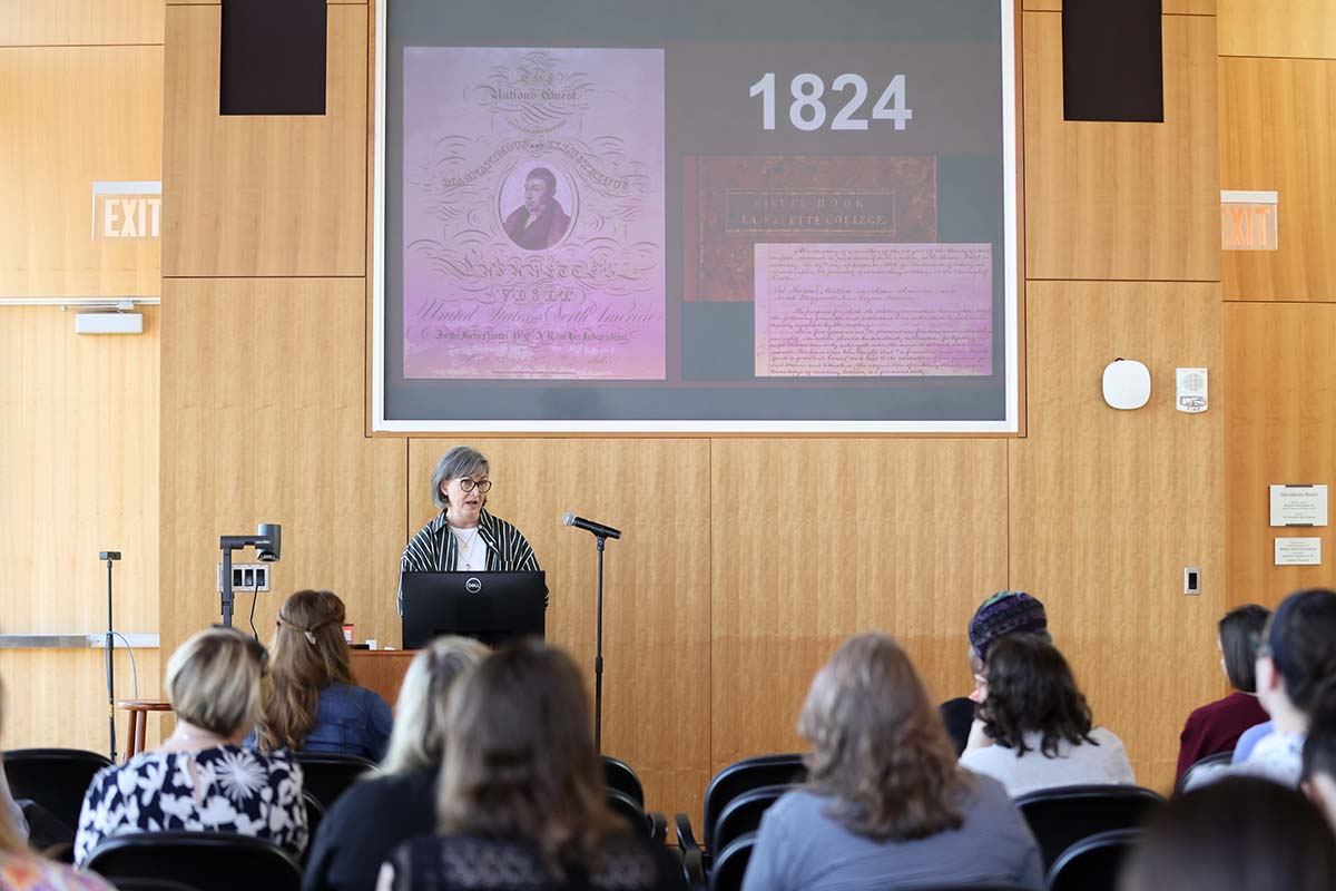 Elaine Stomber stands in front of crowd and a screen showing Lafayette's letters