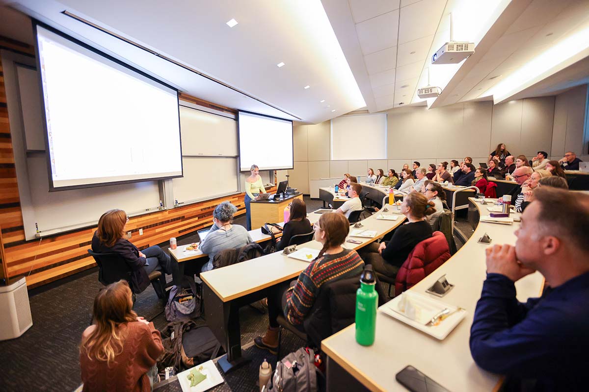 Attendees sit in a lecture hall listening to a student presentation. 
