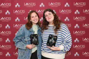 Hope Basaman and Katie Simpson stand together holding Landis plaques against a Landis logo background.