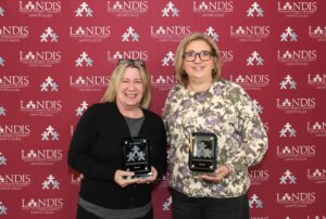 Two recipients of the Landis community engagement awards stand, holding their awards in front of a Landis logo background