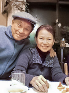 Dieu and his wife, Marie, are seated at a table and smiling at the camera. Dieu is wearing a gray hat.