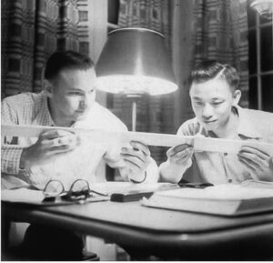 Dieu is pictured with a classmate. They are seated at a table and are holding a long ruler. There is a standing lamp behind them.