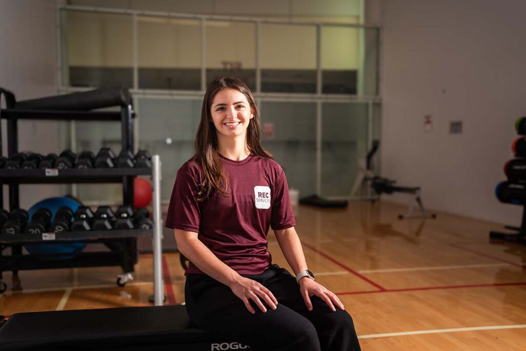 Gia Mazza sits in a weight room wearing a Rec Services shirt. 