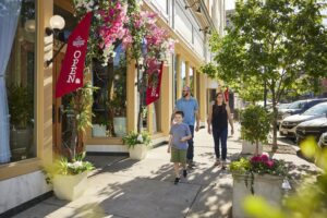 Two adults and a child walk down the sidewalk in front of Meli European Taverna which sports pink flowers over its sign and red "Open" flags by the door.