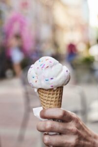 A scoop of vanilla ice cream with sprinkles in a sugar cone is held with Bank Street Alley in the background.