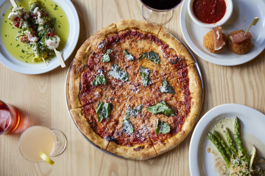 A full pizza, dotted with basil, sits on a table with several appetizers and beverages.