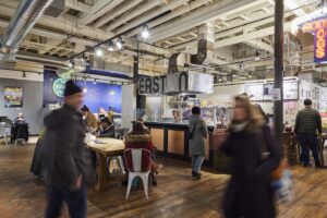 A view inside Easton Public Market as passersby walk past Rod's Dogs and Modern Crumb, with blurs indicating the motion of customers.