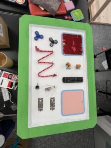 Green trim sensory board features fidget spinners, flip sequins, wheels, and everyday tactile objects.