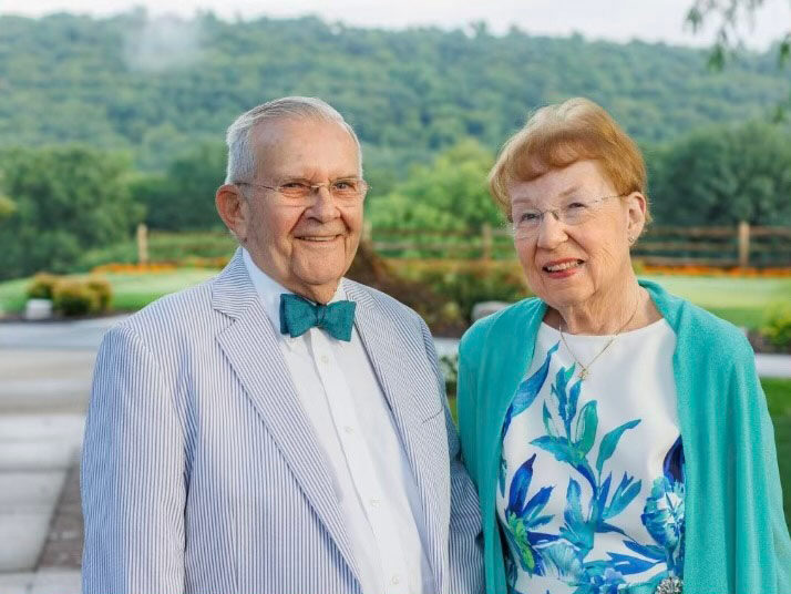 Gordon and Ruth Macdonald are smiling at the camera. Gordon is wearing a green bow tie. Ruth is wearing a green sweater.