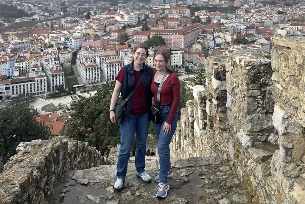Madison Horvath ’26 is shown at right with Maya Maynard ’27 during the 2024 interim trip to Portugal. They stand on stone steps with views of the city below the,