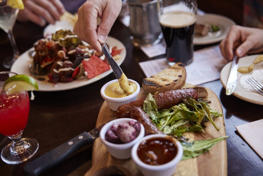 A charcuterie board and nachos are pictured on a table as several diners dig in and spread mustard.