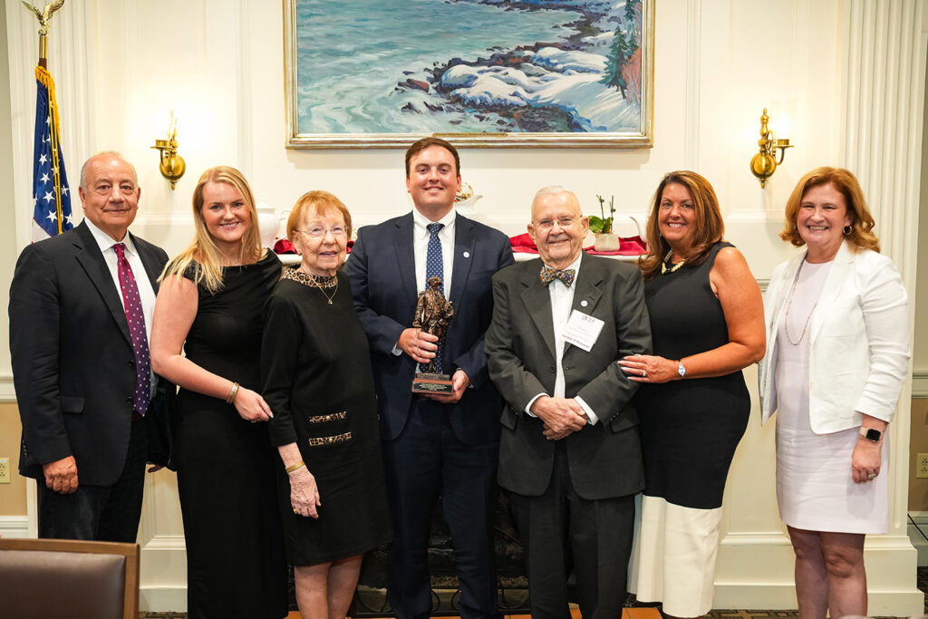 Outgoing Board Chair Bob Sell and President Nicole Hurd pose with five alumni. One alum is holding a mini statue of Marquis de Lafayette.
