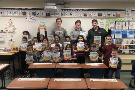 Players stand behind a group of second graders, each holding a book as part of the 2nd & 7 program.
