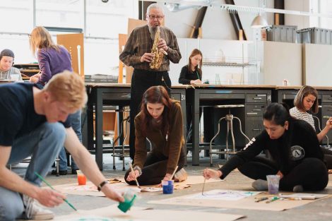 Gary Hassay stands in the background and plays saxophone while students sit on the floor and paint