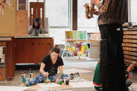 Gary Hassay stands in the foreground and plays saxophone while a student sits on the floor and paints
