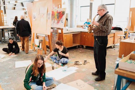 Gary Hassay stands to the right of a student who sits cross-legged and paints
