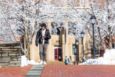 A student looks at his smart phone while walking on a brick walkway after snowfall.