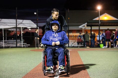Baseball player helps push a man in a wheel chair to first base.