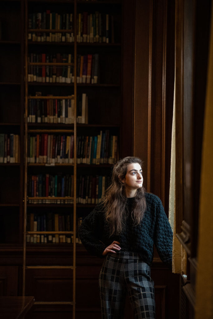 Victoria Puglia ’21 in front of a bookshelf of Kirby Library; she looks out a window with her hand on her hip.