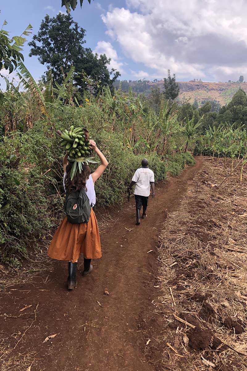 Victoria Puglia ’21, facing away from the camera, carries bananas above her head; she is following a person on a dirt path, with greenery and blue skies surrounding her. 