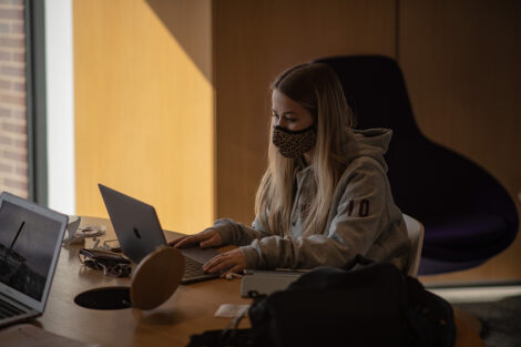 A masked student uses their laptop at a table inside of Rockwell Integrated Sciences Center.