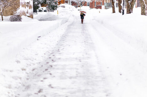 A student walks away from the camera, down a snow-covered path. They are carrying an umbrella