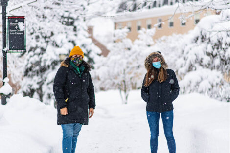 Two masked students stand socially distant, surrounded by snow on the ground and trees.