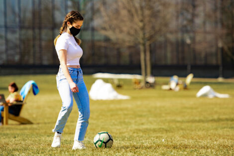 A masked student kicks a soccer ball on the Quad. Skillman Library is behind them.