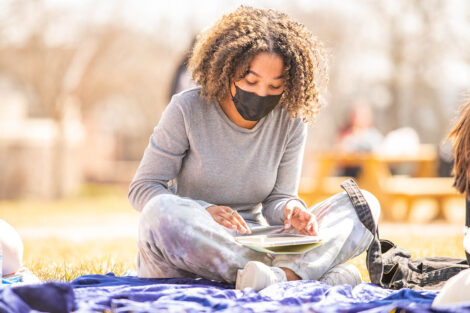 A masked student reads a book, sitting on a blanket on the Quad.